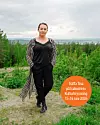 En kvinna står på en grusstig med grön skog och molnig himmel i bakgrunden.