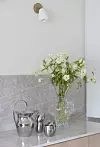 Vita blommor i glasvas på köksbänk med metallkannor och kopp under vägglampa.