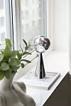 Polerad metallskulptur på en bok vid ett fönster med en grön växt bredvid.