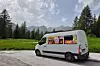 Vit skåpbil parkerad vid en väg med skog och berg bakom.