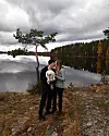 Ett par står vid en sjö och håller en vit katt, med höstskog och molnig himmel i bakgrunden.