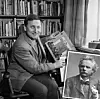 En man sitter framför en bokhylla och håller upp en bok bredvid ett porträttfotografi.