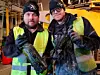 Två män i reflexvästar och mössor håller upp mörka, leriga föremål inomhus i en industriell miljö.