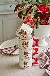 Fyra vita muggar med röda julmotiv staplade på ett bord intill servetter och en blomsterarrangemang.