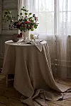 Ett bord med beige duk, blomsterarrangemang och porslin vid ett solbelyst fönster.