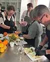 Flera personer lagar mat runt en köksbänk med frukt, grönsaker och glas i ett ljust kök.
