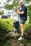 Två män står på en stenig stig i ett grönt naturområde, med en rullstol mellan dem.