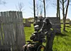 Rullstolsanvändare i paintballmask pekar med markör på en gräsplan med två spelare bakom ett träskydd.