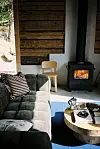 Rustik stuginteriör med grön soffa, vedkamin och runt träbord vid fönster.