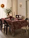 Dukat matbord med blommig duk, ljusstakar och vas i en ljus matsal med flera stolar.