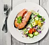 En vit tallrik med laxfilé, grönsaker och citron på ett grått träbord bredvid bestick.