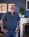 Äldre man står i ett vardagsrum framför en öppen spis och en golvlampa.