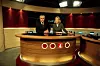 Två programledare vid ett nyhetsbord i en tv-studio med röda väggar och skärmar.