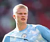 En blond fotbollsspelare i ljusblå tröja står på planen med oskarp publik bakom sig.