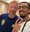 Två män tar en selfie inomhus, den ena i blå tröja med svensk fotbollslogga och den andra gör fredstecken.