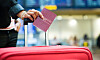 En hand håller upp ett pass bredvid en röd resväska i en flygplatsterminal.