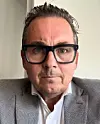 En man i glasögon och grå kostym sitter framför en ljus vägg och tittar in i kameran.