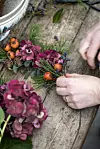 Händer binder en blomsterdekoration med lila blommor, grönt och små bär på ett träbord.