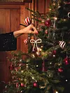 En hand placerar en randig julgranskula på en julgran med röda kulor och ljusslinga.