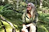 Kvinna sitter på mossig mark i skogen bland ormbunkar och nedfallna grenar.