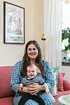 En kvinna sitter i en rosa soffa och håller ett leende barn i knät i ett ljust vardagsrum.