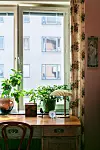 Trämöbel framför ett fönster med krukväxter, bordslampa och liten klocka i ett solbelyst rum.