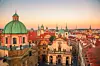 Panoramavy över Prag med gröna kupoler, kyrktorn och tätt bebyggda röda tak vid solnedgång.