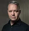 En man i svart skjorta poserar framför en mörk, texturerad bakgrund.