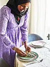 Kvinna i lavendellila klänning skär grönsaker på en färgglad tallrik vid ett dukat bord.