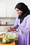 En kvinna i lila klänning och svart huvudduk blandar sallad vid ett köksbord i ett ljust kök.