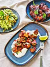 Tre blå tallrikar med sallad, grillat kött och lime på ett ljust bord med en gaffel.