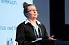 Kvinna i svart kavaj står vid ett podium och talar framför en presentationsskärm.