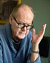 Peter Haber i glasögon och blå tröja som sitter inomhus med handen lyft vid ansiktet.