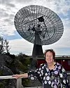 Stort radioteleskop mot molnig himmel med en kvinna vid ett räcke i förgrunden.