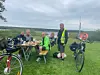 Fem personer sitter och står vid ett bord på en gräsmatta med cyklar och en sjö i bakgrunden.