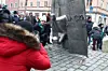 Flera personer står runt en staty i en stadsmiljö med blommor och ett tänt ljus vid sockeln.