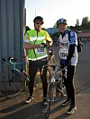 Två cyklister står bredvid sina cyklar utomhus i solen, med hjälm och nummerlappar.