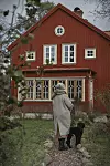 Person med svart hund på grusgång framför rött hus med vita fönster.