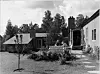 Svart-hvitt bilde av et hus med gårdsplass, trær og en liten hage.