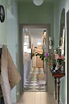 En smal, ljusgrön hall med jacka, speglar och blomsterarrangemang som leder mot ett kök i bakgrunden.