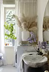Ljust badrum med pampasgräs, blommor och rund marmorvask intill ett fönster.
