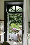 Uteplats med bord, blommor och en person som sitter vid en fransk balkong mot en grön trädgård.