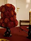 Röd blomsterarrangemang i svart vas på vinrött dukat bord med ljus.