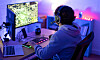 Person med headset sitter vid skrivbord med datorer och skärmar i ett rum med blå och lila belysning.