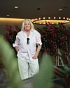 Mia Parnevik i ljusrosa skjorta och ljusa byxor. Hon ler mot kameran och har ett par solglasögon hängandes i skjortan. Växter syns i förgrunden.