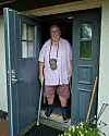 Man i rosa skjorta står i en öppen ytterdörr med två käppar i händerna.