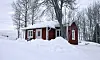 Röd stuga med vita detaljer står i djup snö bland kala träd under en molnig vinterdag.