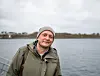 Mann i grønn jakke og grå lue står ved vannet på en overskyet dag.
