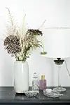 Vas med torkade blommor på ett bord bredvid en vit bordslampa och små dekorativa föremål.