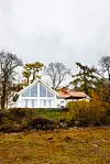 Vitt hus med stora fönster vid skogskant, omgivet av höstfärger och buskar under grå himmel.